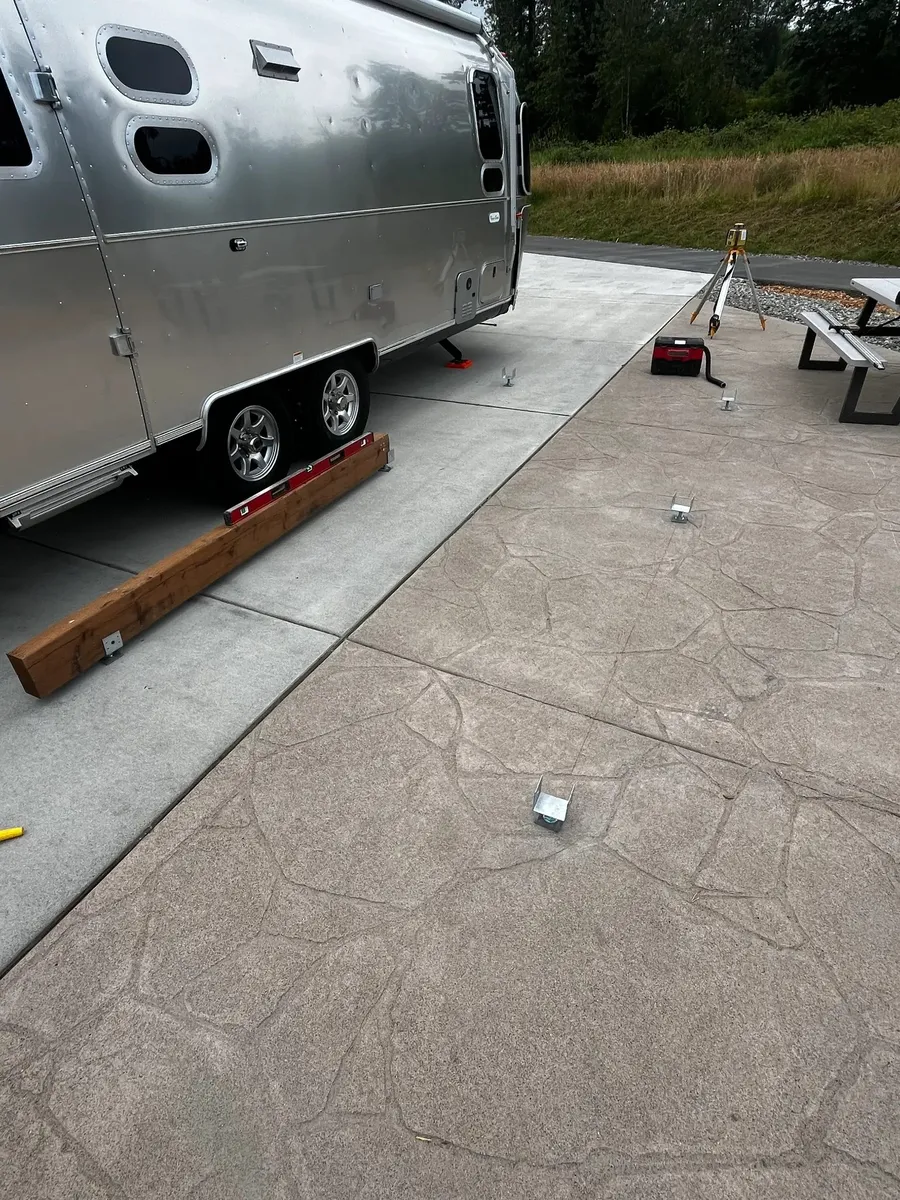 Deck anchor brackets laid out on stamped concrete pad beside an Airstream trailer with a laser level set up for precise placement at Angel of the Winds RV Resort